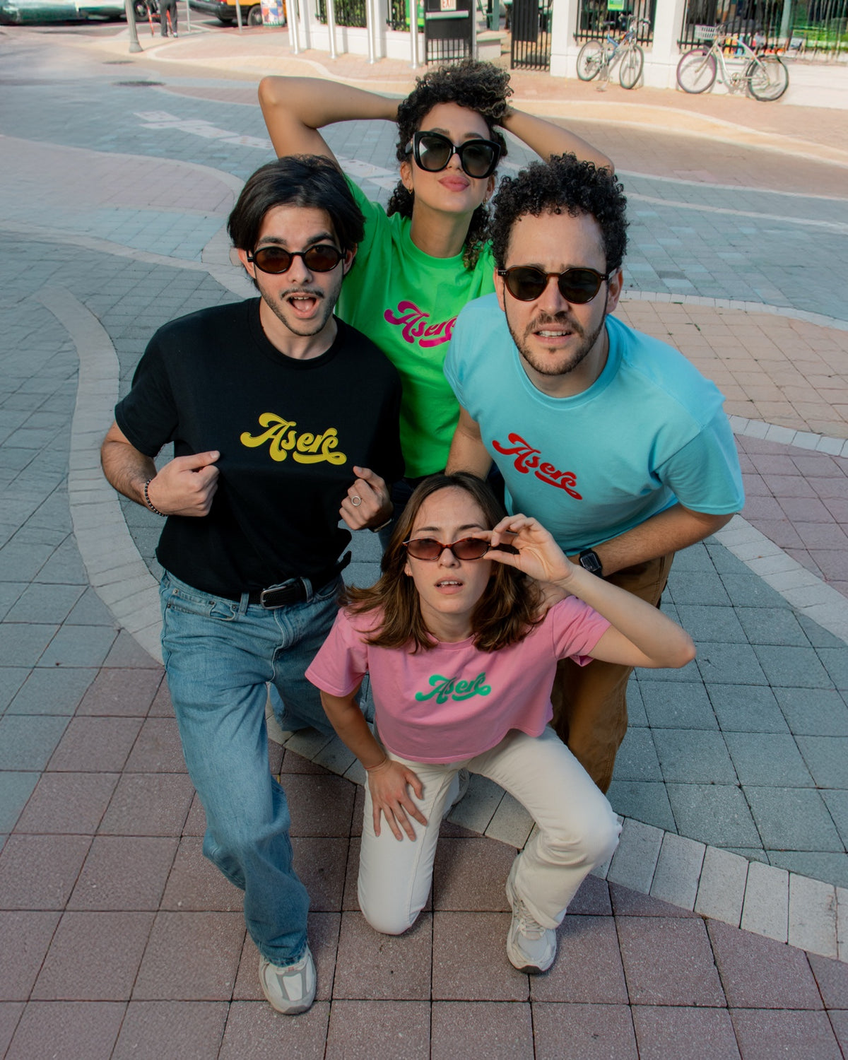 Cuatro personas luciendo camisetas Asere en negro, verde, azul y rosa.